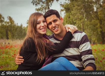 Young couple sitting on the grass in a field of red poppies and smiling at the camera.