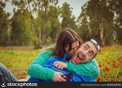 Young couple sitting on the grass in a field of red poppies. The girl kiss the guy while he has a silly smile.