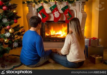 Young couple sitting under Christmas tree and looking at burning fireplace