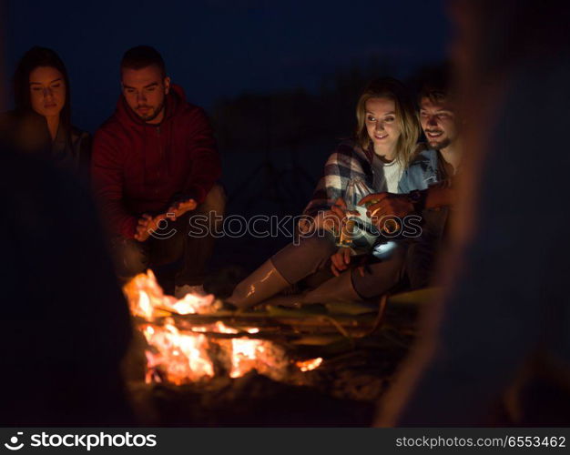 Young Couple Sitting with friends Around Campfire on The Beach At Night drinking beer. Couple enjoying with friends at night on the beach