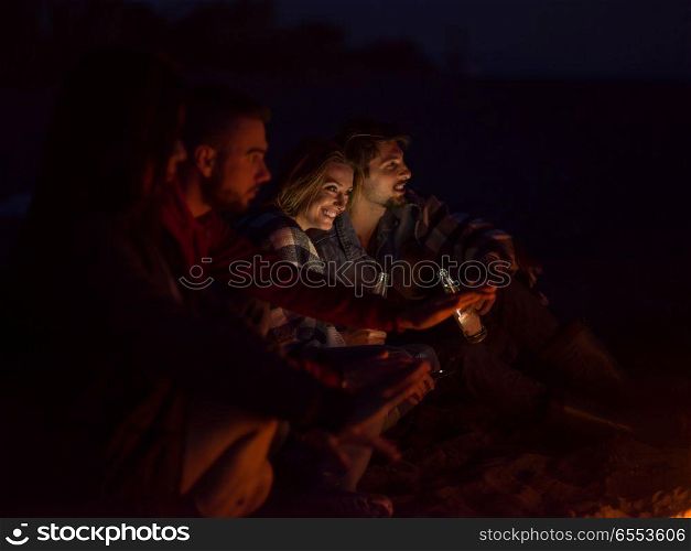 Young Couple Sitting with friends Around Campfire on The Beach At Night drinking beer. Couple enjoying with friends at night on the beach