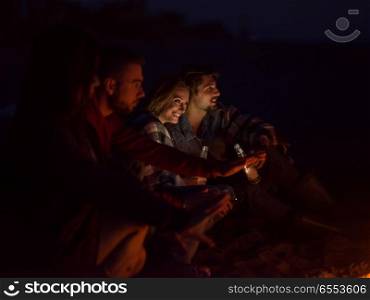 Young Couple Sitting with friends Around Campfire on The Beach At Night drinking beer. Couple enjoying with friends at night on the beach