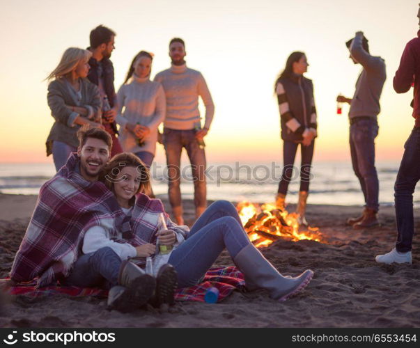 Young Couple Sitting with friends Around Campfire on The Beach At sunset drinking beer. Couple enjoying with friends at sunset on the beach