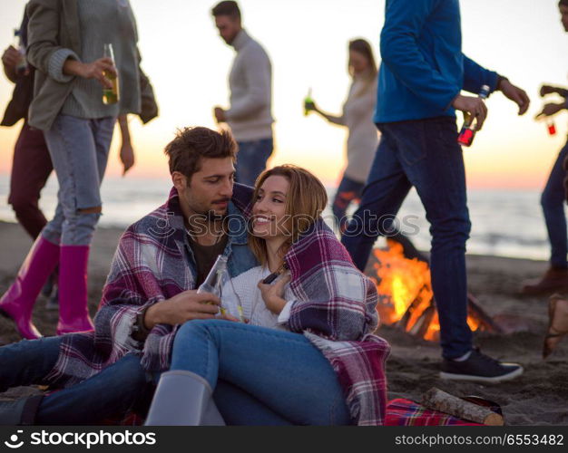 Young Couple Sitting with friends Around Campfire on The Beach At sunset drinking beer. Couple enjoying with friends at sunset on the beach