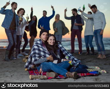 Young Couple Sitting with friends Around Campfire on The Beach At sunset drinking beer. Couple enjoying with friends at sunset on the beach