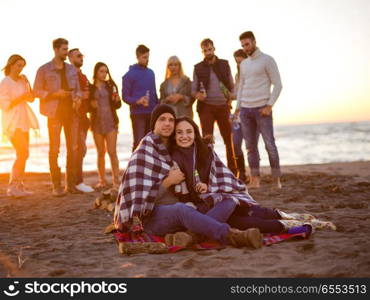 Young Couple Sitting with friends Around Campfire on The Beach At sunset drinking beer. Couple enjoying with friends at sunset on the beach