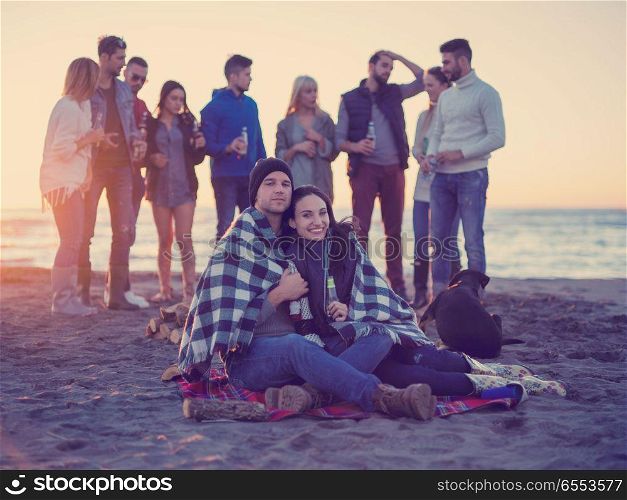 Young Couple Sitting with friends Around Campfire on The Beach At sunset drinking beer. Couple enjoying with friends at sunset on the beach