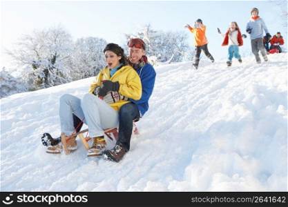 Young Couple Sledging Down Hill With Family Watching