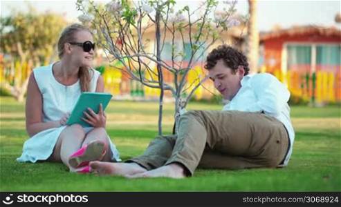 Young couple spending free time on the green lawn. Woman using a touchpad, man reading a book