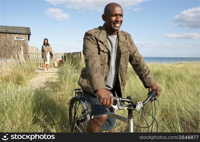 Young Couple Standing By Beach Hut Amongst Dunes With Man On Mountain Bike