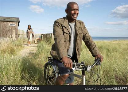 Young Couple Standing By Beach Hut Amongst Dunes With Man On Mountain Bike