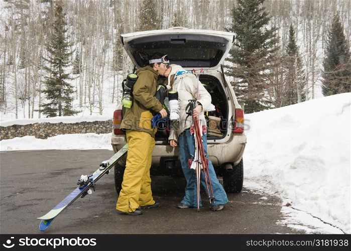 Young couple standing by vehicle with ski equipment kissing.