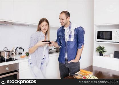 young couple standing kitchen using mobile phone while cooking food