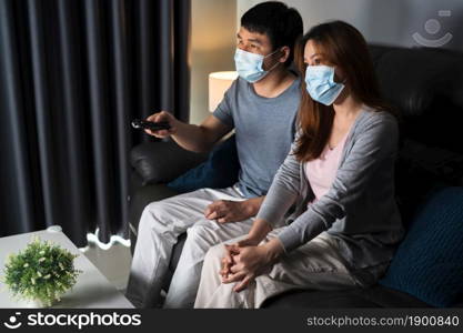young couple watching TV on sofa and wearing medical mask to protect coronavirus (Covid-19)