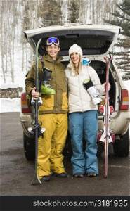 Young couple wearing winter clothes standing by automobile with ski equipment smiling.