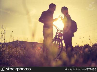 Young couple with bike are standing on a field and doing a gesture, sundown scenery