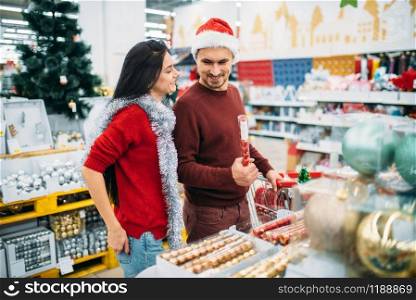 Young couple with cart in department of holiday decorations in supermarket, family tradition. December shopping of new year or christmas goods. Young couple with cart, department of decorations