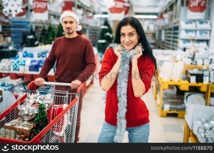 Young couple with cart in department of holiday decorations in supermarket, family tradition. December shopping of new year or christmas goods