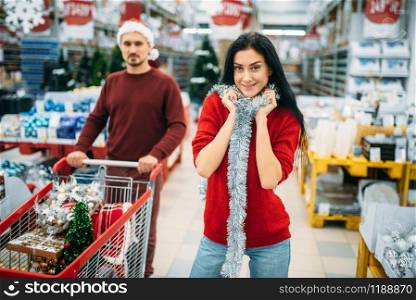 Young couple with cart in department of holiday decorations in supermarket, family tradition. December shopping of new year or christmas goods