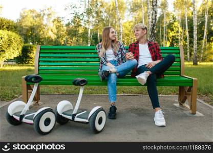 Young couple with gyroboard sitting on the bench in park. Outdoor recreation with electric gyro board. Transport with balance technology. Couple with gyroboard sitting on the bench in park
