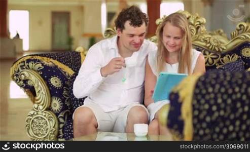 Young couple with touchpad sitting on the sofa in the hotel lobby. Woman showing a man something on pad while he having tea