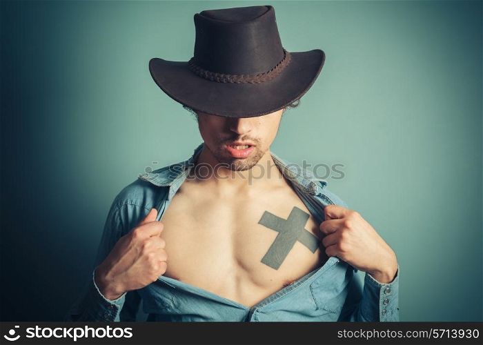 Young cowboy is unbuttoning his shirt to reveal a tattoo on his chest