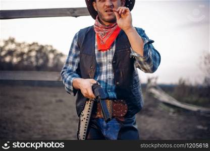 Young cowboy with gun poses against horse corral, western. Vintage male person, retro american fashion in wild west style