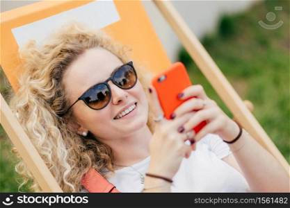 Young curly blonde woman lies in hammock, enjoys summer day, bloggs and chats via smart phone, wears trendy shades, has positive smile, recreats during holidays. People and lifestyle concept.