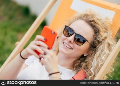 Young curly blonde woman lies in hammock, enjoys summer day, bloggs and chats via smart phone, wears trendy shades, has positive smile, recreats during holidays. People and lifestyle concept.