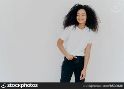 Young curly haired lady has slim figure, wears white t shirt and jeans, looks aside with happy expression, shows healthy teeth, notices funny scene on right side, isolated on white background
