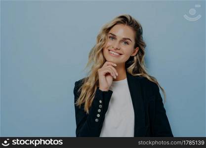 Young cute blonde woman touching with hand her chin, looking straight while smiling with big smile, making joyful and positive expression, posing isolated on blue background with copy space for text. Young blonde woman with hand on her chin smiling at camera