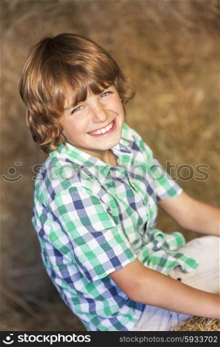 Young cute happy smiling boy wearing a plaid shirt sitting smiling on bales of hay or straw in a barn