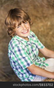 Young cute happy smiling boy wearing a plaid shirt sitting smiling on bales of hay or straw in a barn