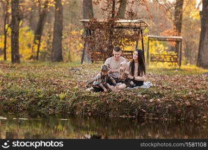 young dad and mom with baby girl and little son having fun on blanket near pond in autumn park on sunny day. happy family concept. mother&rsquo;s, father&rsquo;s, baby&rsquo;s day. young dad and mom with baby girl and little son having fun on blanket near pond in autumn park on sunny day. happy family concept. mother&rsquo;s, father&rsquo;s, baby&rsquo;s day.