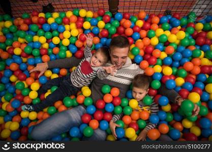 Young dad plays with kids at pool with colorful balls in a children&rsquo;s playroom