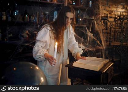 Young demonic woman with candle reads book of spells, demons casting out. Exorcism, mystery paranormal ritual, dark religion, night horror, potions on shelf on background. Demonic woman with candle reads book of spells