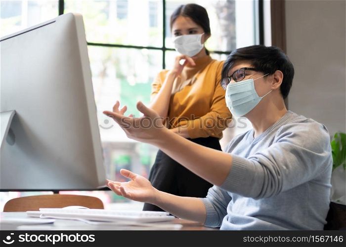 Young Designers in protective face mask for protect pandemic virus working on computer monitor together. Asian Businessman and businesswoman discussing and consulting business job in meeting at office