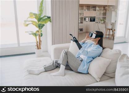 Young disabled attractive european woman is sitting in living room on couch. Girl with cyber arm in vr glasses. Handicapped person gets rehabilitation at home. Excited lady touches 3d vision.. Disabled european girl sitting on couch. Woman with cyber arm in vr glasses gets rehabilitation.