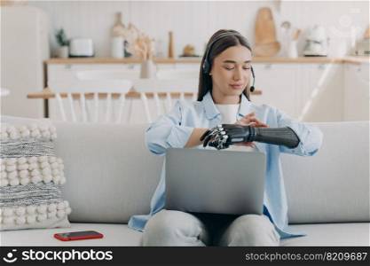 Young disabled girl freelance employee in headset working at laptop online, sitting on sofa at home, setting her bionic prosthetic arm. People with disabilities and daily routine concept.. Young disabled girl wearing headset working at laptop at home, setting her bionic prosthetic arm