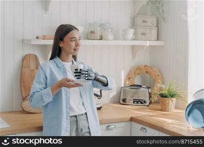 Young disabled girl holding mug by bionic prosthetic arm, standing in cozy kitchen. Female with disability holds cup by artificial robotic hand, drinking morning coffee, enjoying domestic routine.. Young disabled girl holding mug by bionic prosthetic arm, enjoying domestic routine in cozy kitchen