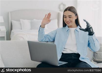 Young disabled woman has conference online on laptop. Remote work and freelance for handicapped girl. Attractive woman with artificial arm at home. Modern bionic prosthesis, technology concept.. Young disabled woman has conference online on laptop. Remote work for handicapped girl.