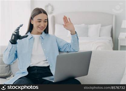 Young disabled woman has conference online on laptop. Remote work and freelance for handicapped girl. Attractive woman with artificial arm at home. Modern bionic prosthesis, technology concept.. Young disabled woman has conference online on laptop. Remote work for handicapped girl.