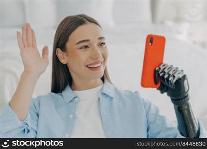 Young disabled woman has video call on phone. European girl is holding the mobile phone with bionic artificial arm and taking selfie. Happy caucasian woman with artificial limb at home.. Young disabled woman has video call on phone. European girl is holding phone and taking selfie.