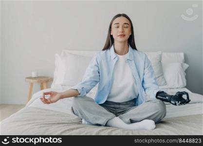 Young disabled woman is meditating in lotus pose on bed in her bedroom. Handicapped caucasian girl has sensored bionic prosthesis with her eyes closed. Myoelectric artificial limb. Stress relief.. Young disabled woman is meditating in lotus pose on bed in her bedroom. Myoelectric artificial limb.
