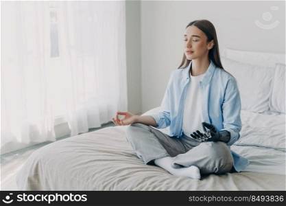 Young disabled woman is practicing yoga in lotus pose on bed in her bedroom. Stress relief at home. Attractive european girl has bionic cyber prosthesis. Modern artificial limb of high technology.. Young disabled woman is practicing yoga in lotus pose on bed in her bedroom. Stress relief at home.