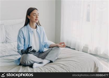 Young disabled woman is practicing yoga in lotus pose on bed in her bedroom. Stress relief at home. Attractive european girl has bionic cyber prosthesis. Modern artificial limb of high technology.. Young disabled woman is practicing yoga in lotus pose on bed in her bedroom. Stress relief at home.