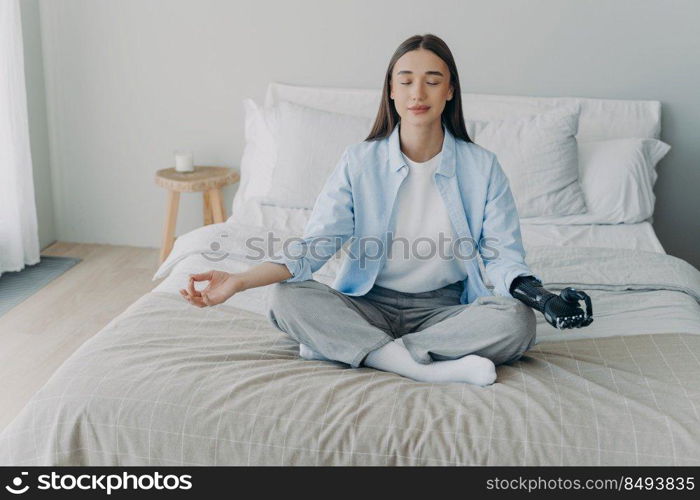 Young disabled woman is sitting in lotus pose on bed with her eyes closed. Attractive european girl has cyber prosthesis. Modern artificial limb. Handicapped woman lifestyle concept.. Young disabled woman is sitting in lotus pose on bed. Handicapped girl lifestyle.