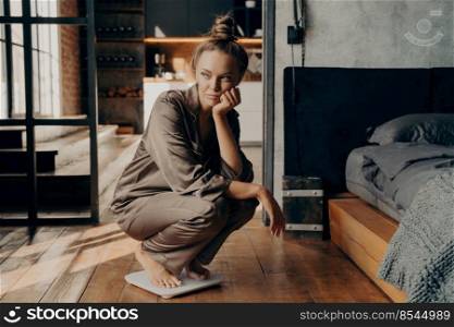 Young dissatisfied unhappy woman crouched over electronic scales in morning after waking up in modern bedroom background, displeased with her weight goals. Dieting concept. Young dissatisfied unhappy woman crouched over electronic scales