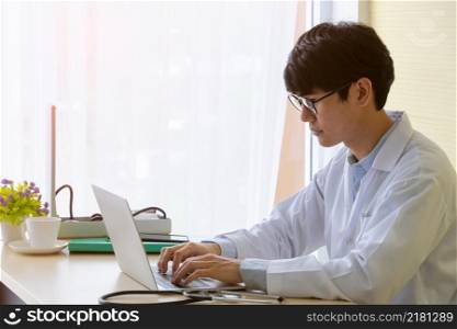 Young doctor physician wearing white uniform and using laptop in hospital office. Male doctor in eyeglasses typing on his computer at desk