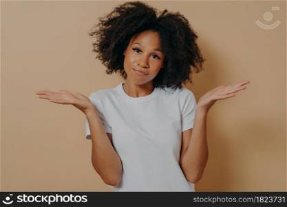 Young doubtful african woman raising palms with hesitation and looking at camera with puzzled face expression, mixed race female being unsure and having some doubts while posing against beige wall. Mixed race female being unsure and having some doubts while posing against beige wall